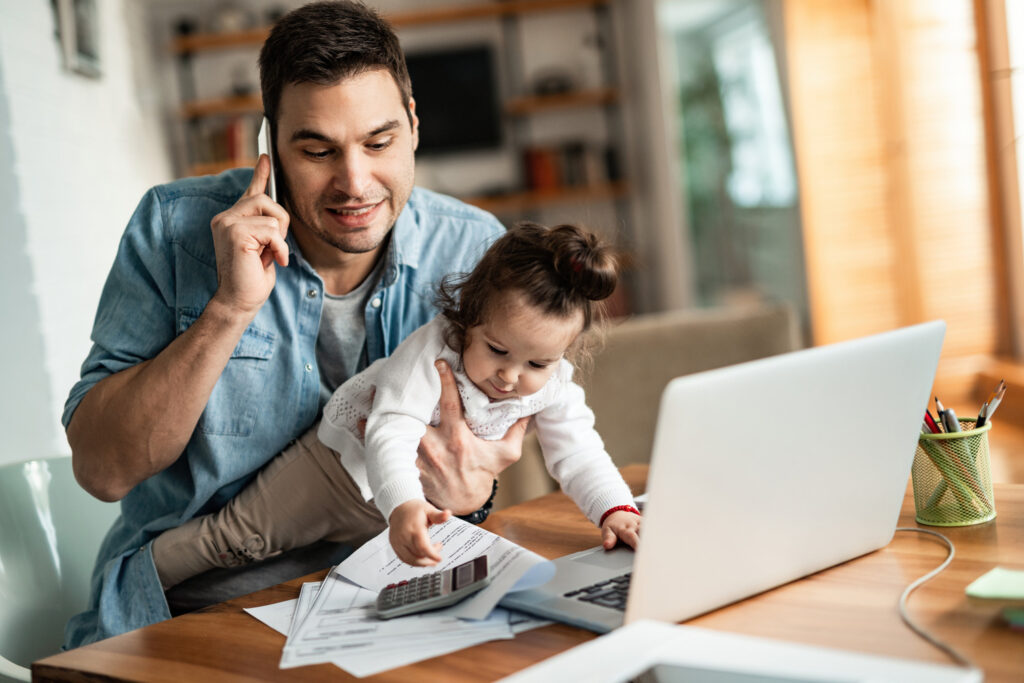 Father holding toddler while trying to manage finances on his phone and laptop